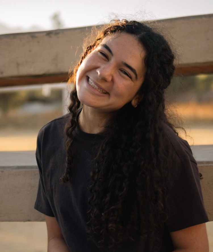 A young woman with long curly dark hair wearing a dark t-shirt tilts her head and smiles outdoors in warm sunlight.