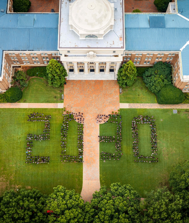 Aerial view of a large group of students standing on a green lawn in front of a domed brick campus building, forming the numbers "2030."