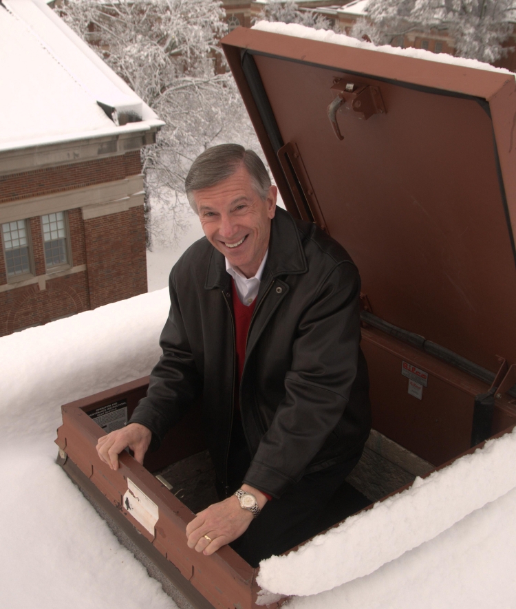 A smiling man in a black leather jacket leans out of a roof hatch onto a snow-covered rooftop overlooking a snow-clad campus.