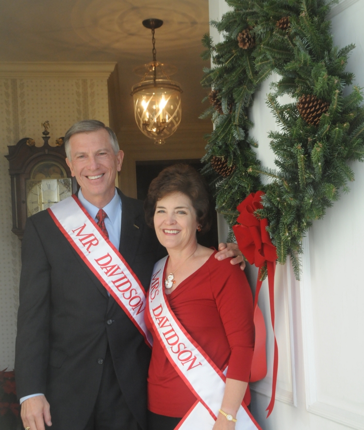 A smiling man in a suit and a woman in a red top stand together in a doorway adorned with a large evergreen wreath, both wearing white sashes reading "MR. DAVIDSON" and "MRS. DAVIDSON."