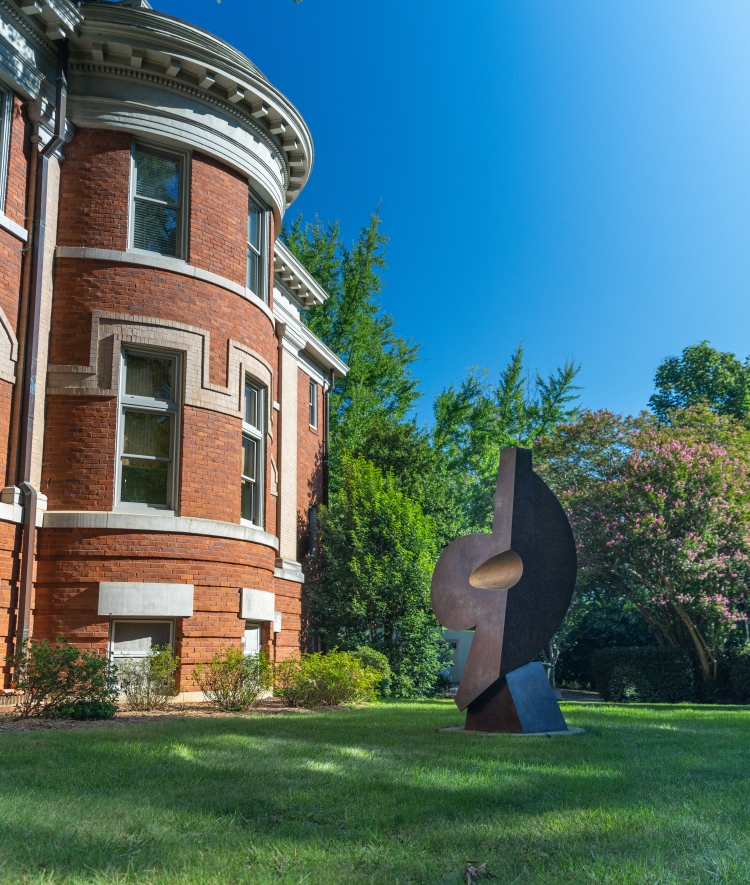 A large red brick campus building with a rounded tower facade, set on a green lawn beside a dark modern abstract sculpture and lush trees under a clear blue sky.