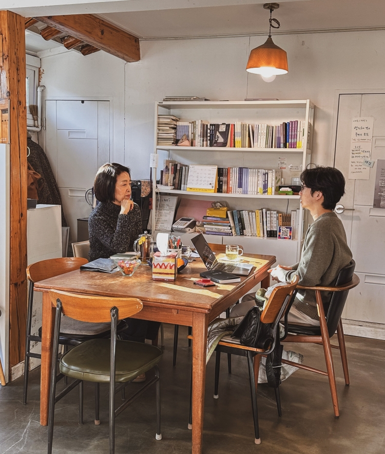 A woman and a young man sit opposite each other at a wooden table in a brightly lit room, engaged in conversation with laptops, snacks, and drinks between them in front of a white bookshelf.