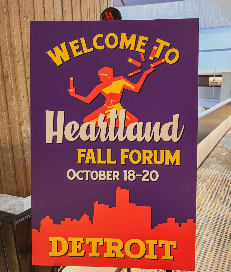 A purple event sign reading "Welcome to Heartland Fall Forum October 18-20 Detroit" displaying a silhouetted figure and skyline graphic.