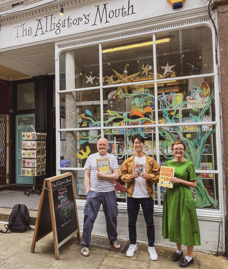 Three people standing outside "The Alligator's Mouth" bookstore holding up books in front of a window decorated with painted trees.