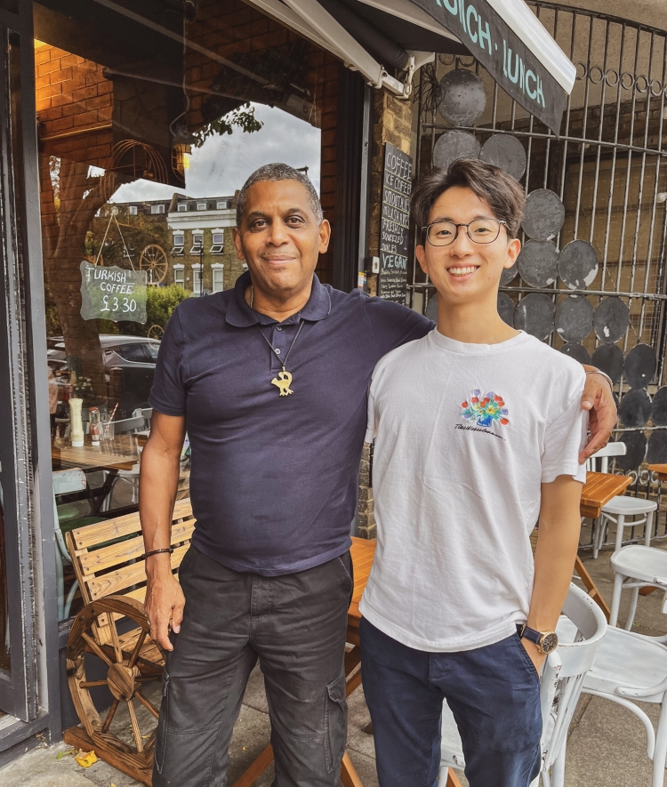 Two men standing side-by-side with their arms around each other outside a café with outdoor seating.