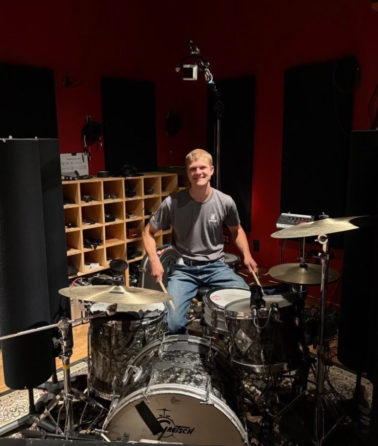 A young man smiles while seated behind a silver sparkle drum set inside a recording studio with acoustic panels and cubby shelving.