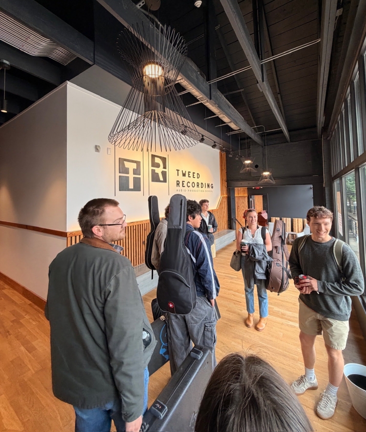 Members of a music group carrying instrument cases gather in the modern lobby of Tweed Recording under a geometric hanging light fixture.