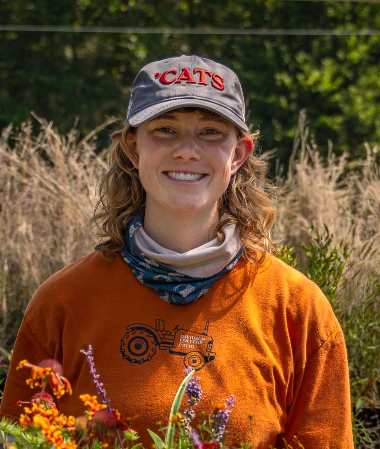 A woman with a 'Cats baseball cap, orange t-shirt, neck gaitor, and shoulder-length blonde curly hair smiles in a greenhouse