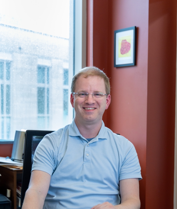 Dr. Thurtle-Schmidt sits at a wooden desk in an office, smiling with glasses and a light blue polo shirt.