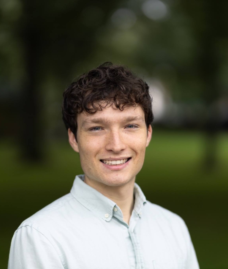 A close-up outdoor portrait of a young man with curly brown hair smiling in front of a soft-focus green background.