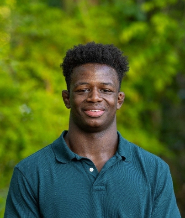 a young Black man wearing a teal collared shirt in front of a blurred green background