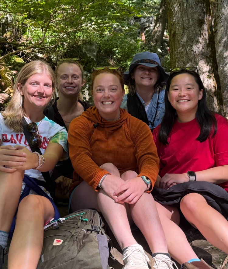 Five smiling friends sit together on large rocks outdoors during a trail hike in a lush, wooded forest.   
