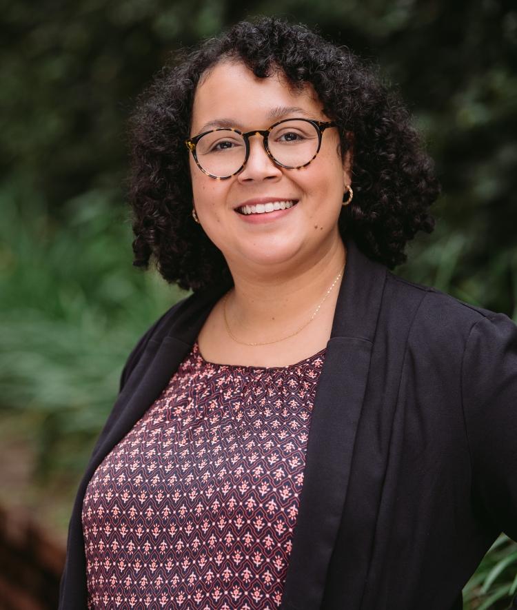 A smiling woman with curly dark hair and round glasses wearing a patterned top and black cardigan, standing outdoors against a soft-focus background of green foliage.