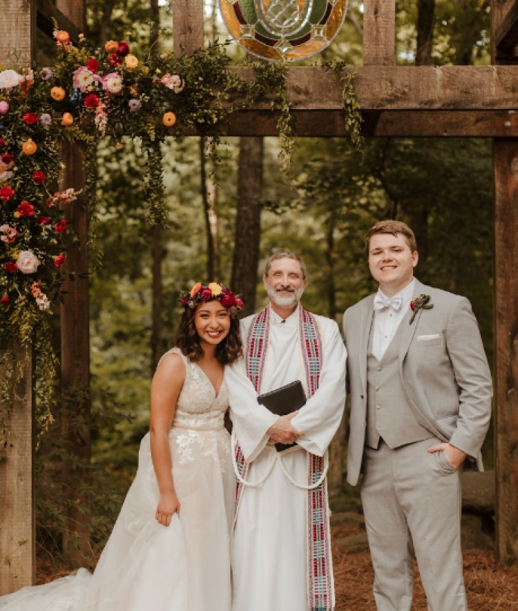 A bride wearing a white lace gown and flower crown stands beside a groom in a light gray suit and a minister beneath a wooden arch adorned with vibrant floral arrangements and stained glass.