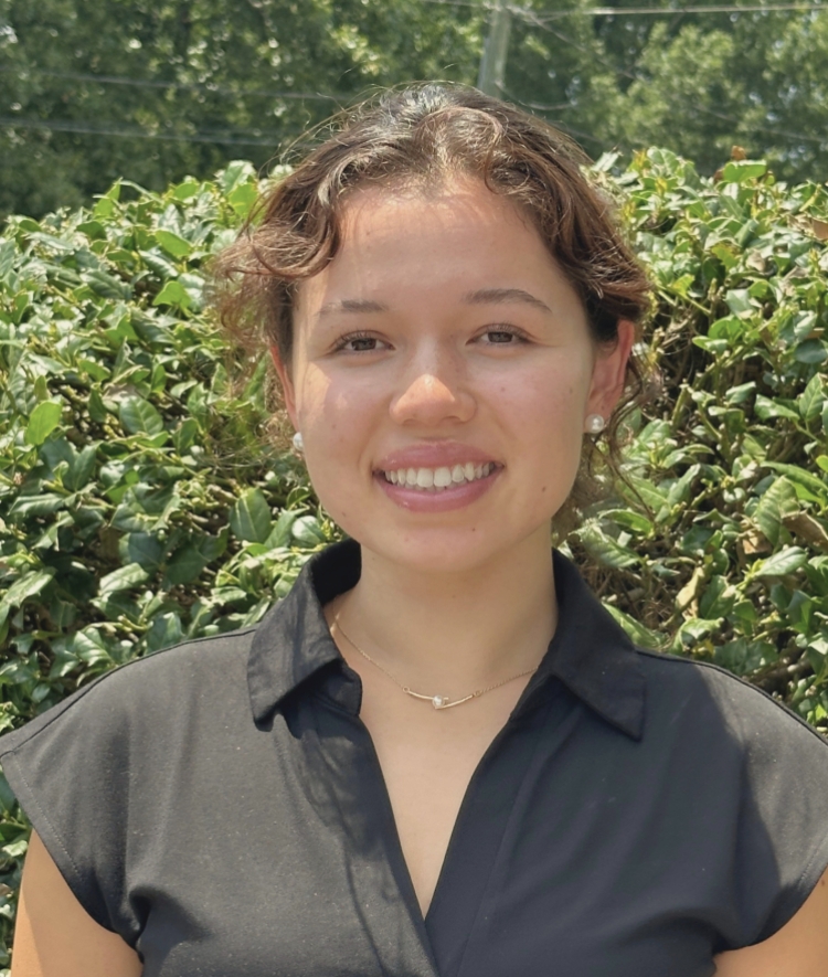 A smiling young woman with brown curly hair pulled back, wearing a black short-sleeved collared shirt and a delicate gold necklace, posed in front of a leafy green hedge.