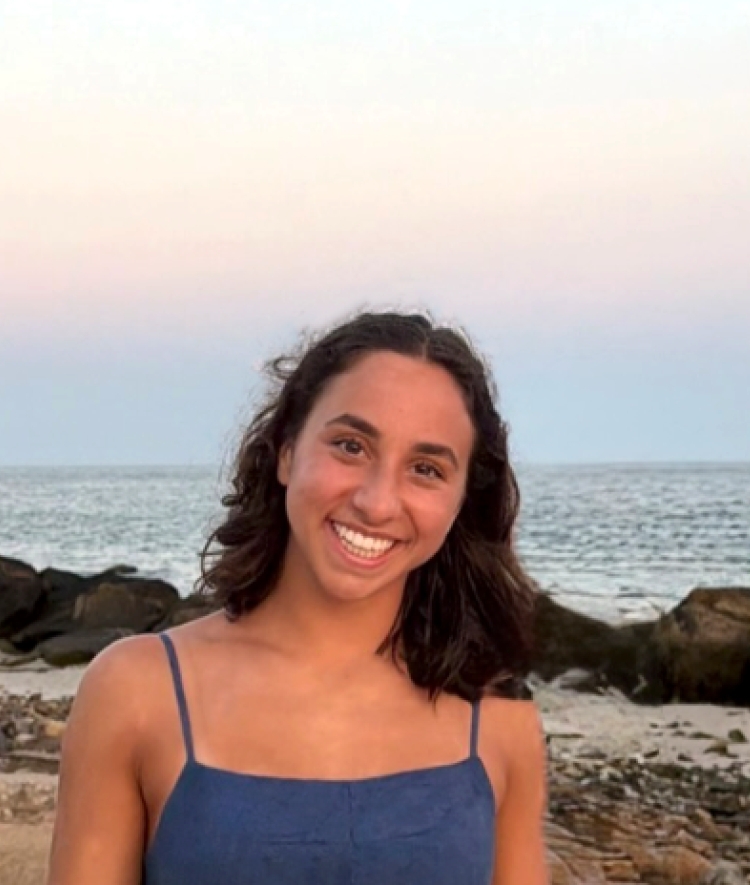 A young woman with shoulder-length dark hair smiles in front of a ocean beach at dusk while wearing a dark blue tank top.