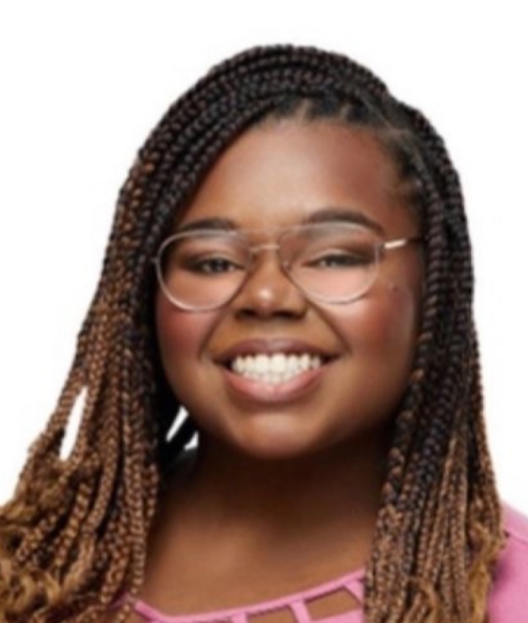 A smiling young Black woman with long braided hair and wire-rimmed glasses wearing a pink top against a plain white background.