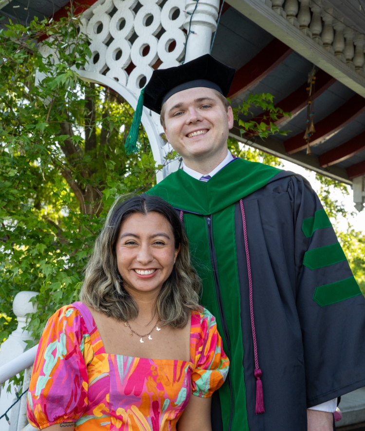 Zach wearing a green and black doctoral graduation gown and mortarboard stands next to Yara dressed in a colorful patterned dress in front of a decorative wooden gazebo.