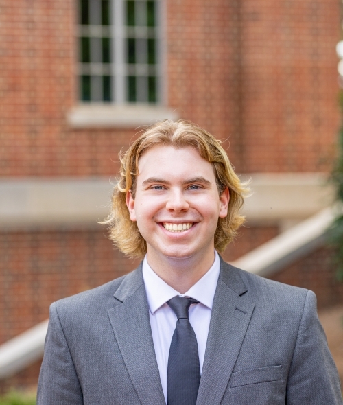 A white male with blonde hair wears a suit and smiles