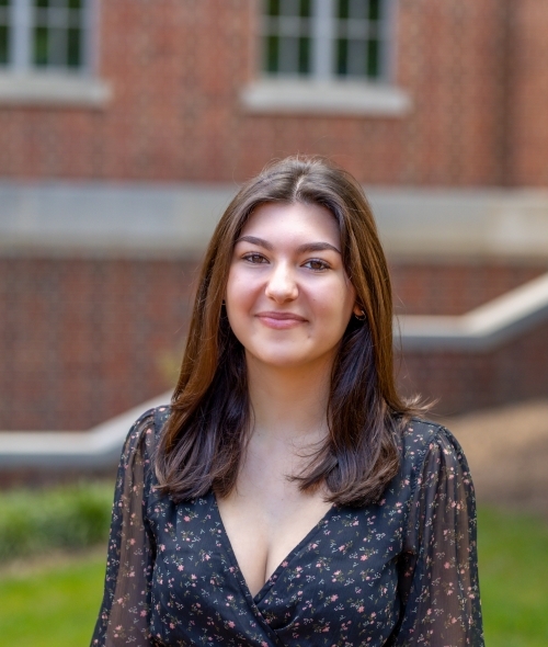 A young woman with dark hair wears a floral black dress and smiles