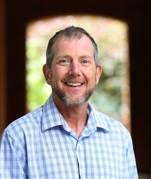 A white male wearing a blue and white gingham shirt smiles and stands with an arch behind him