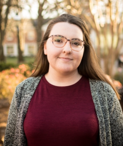 a young white woman wearing a maroon top and grey sweater with glasses and brown hair