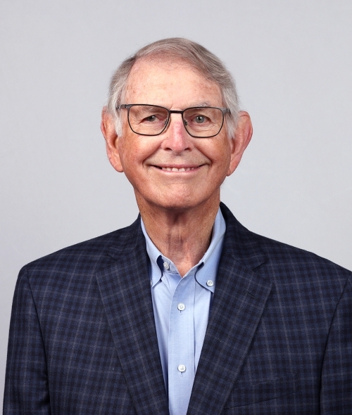 an older white man with grey hair and glasses wearing a blue collared shirt and a plaid blazer
