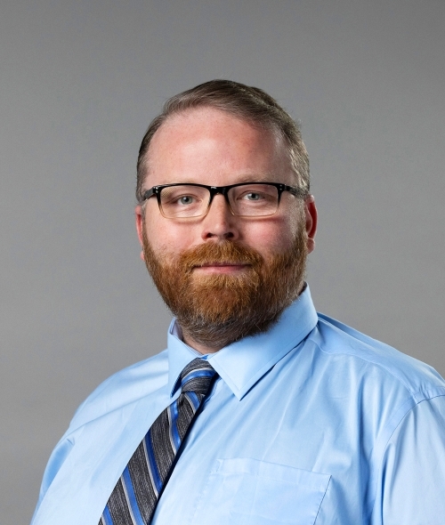a middle aged white man with red hair and facial hair wearing glasses, a blue shirt and a tie