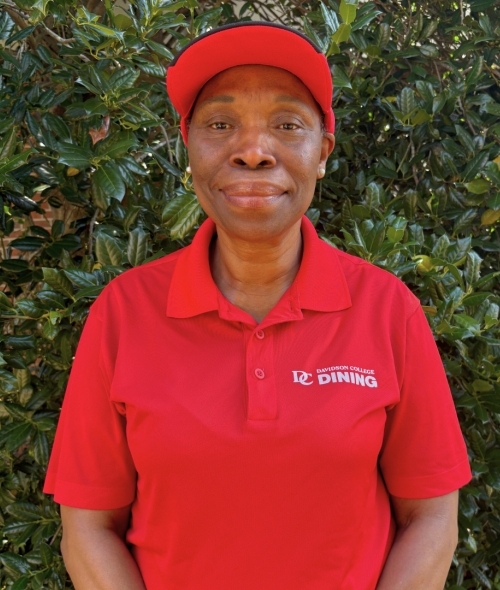 a Black woman wearing a red polo and a red baseball hat in front of a green plant wall