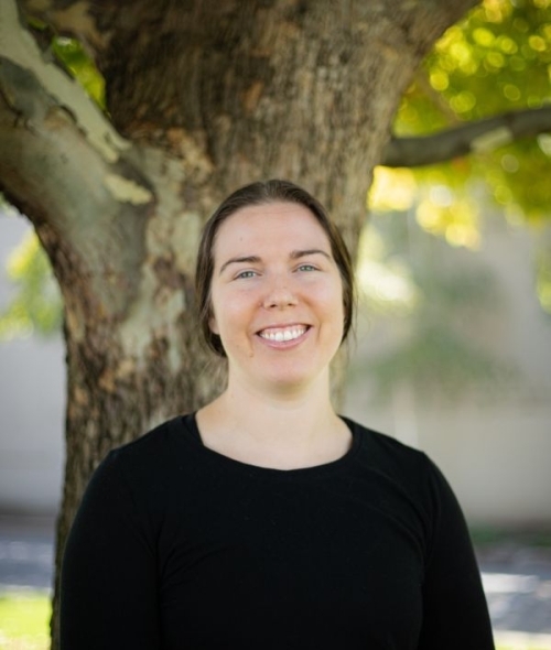 a young white woman with dark hair wearing a black top standing outside in front of a tree and smiling