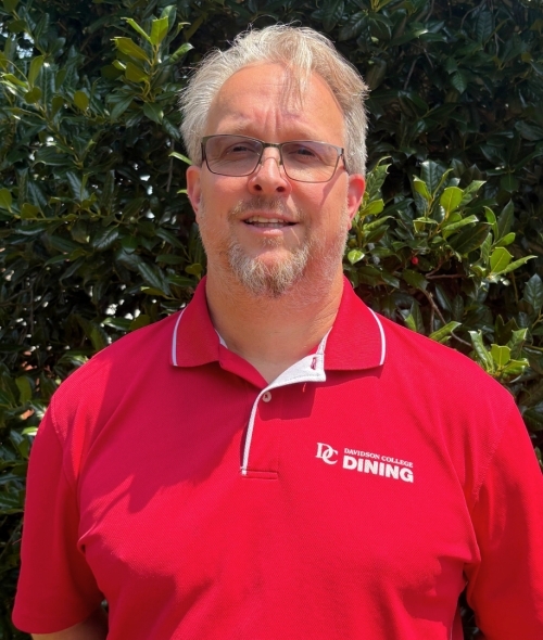 a middle aged white man with white hair and facial hair wearing a red polo shirt on a sunny day