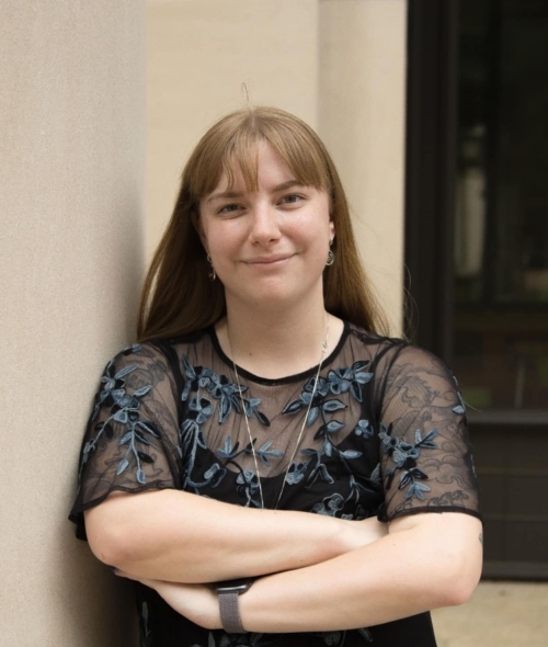 a woman with brown hair and bangs wearing a black top with her arms crossed in front of a stone building