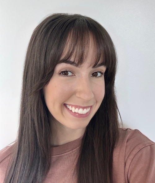 a woman with long straight dark hair smiling wearing a pink top on a grey background