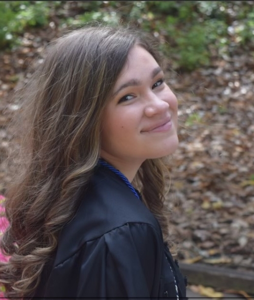 a woman with curly brown hair wearing a black jacket smiling in front of a bed of mulch and flowers