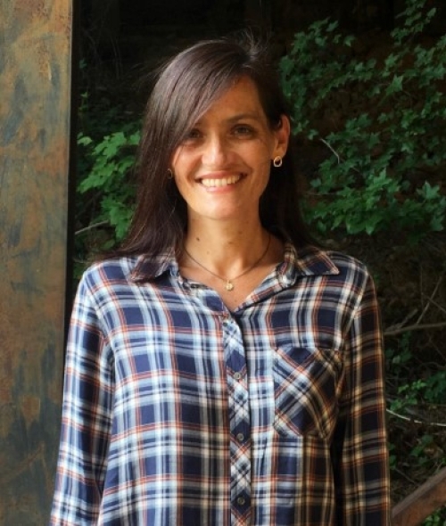 a woman with dark wavy hair wearing a flannel top in front of a green plant