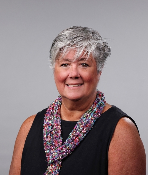 a woman with short grey hair wearing a black top and colorful scarf smiling on a grey background