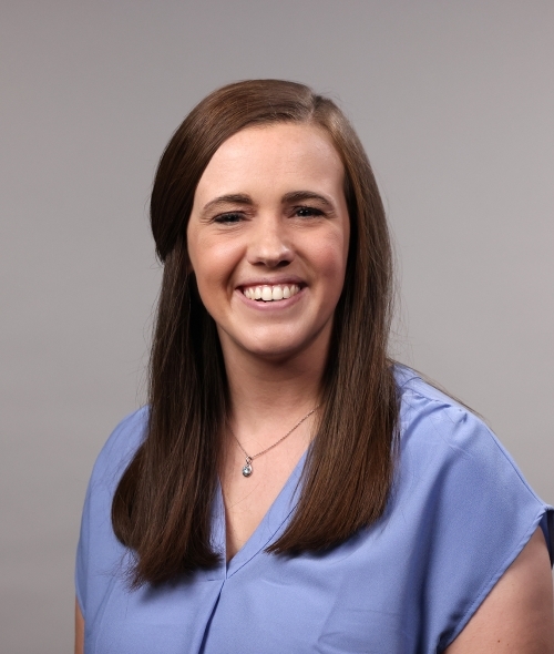 a woman with short brown hair wearing a purple blouse on a grey background