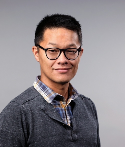 a man with short black shair wearing glasses, a flannel and half zip sweater on a grey background