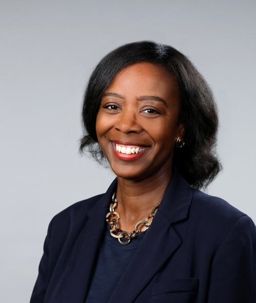 a woman with curly dark hair wearing a blazer and smiling on a grey background