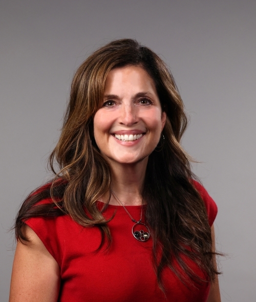a woman with curly brown hair wearing a red top and smiling on a grey background