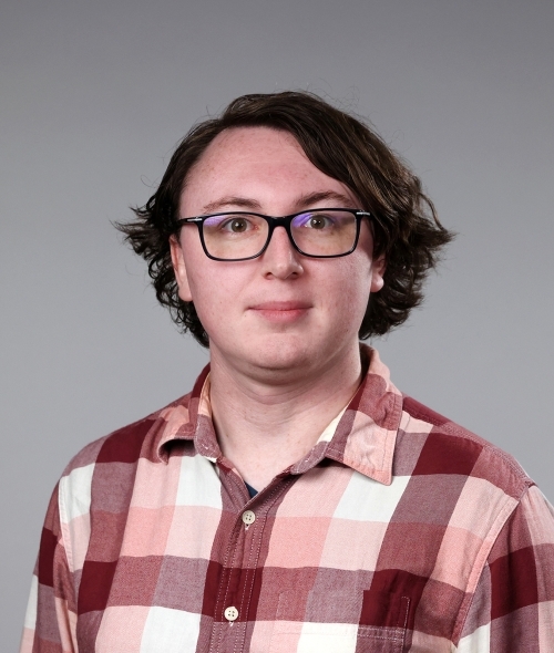 a man with short curly brown hair wearing a red and pink flannel and glasses on a grey background