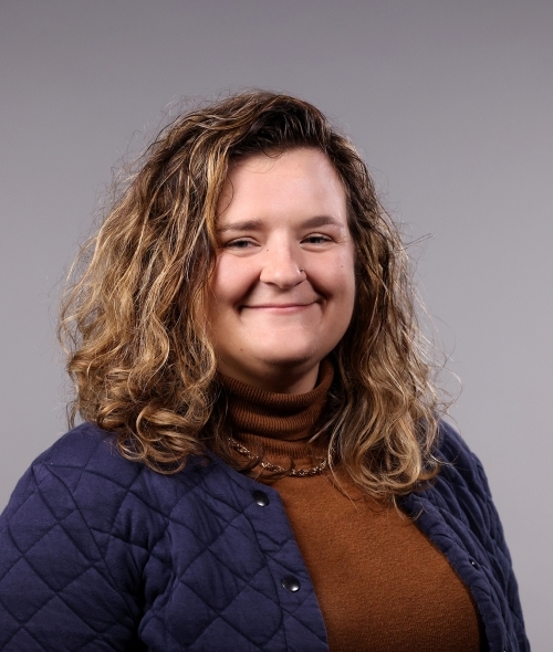 a woman with curly dirty blonde hair wearing a burnt orange turtleneck and quilted jacket on a grey background