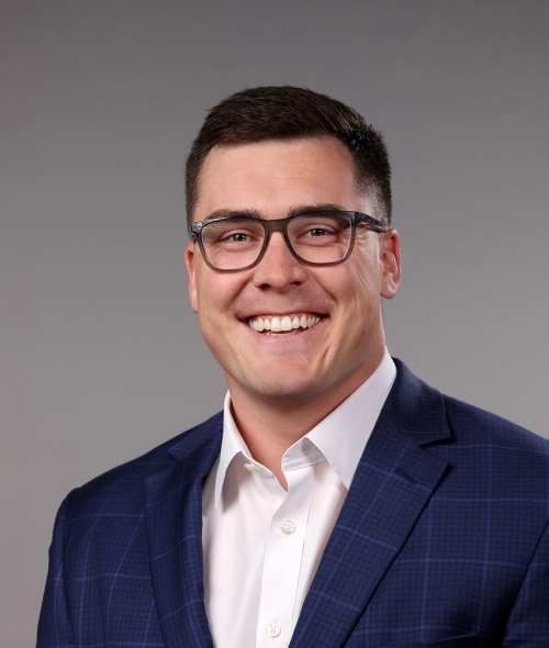 a man with short dark hair wearing a collared shirt and navy blazer and glasses on a grey background