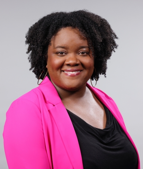 a woman with curly dark hair wearing a black top and pink blazer over a grey background