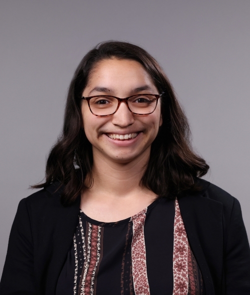 a woman with dark hair wearing red glasses and a dark blouse on a grey background