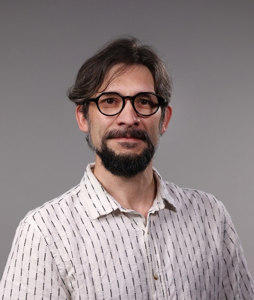 a man with dark hair and facial hair wearing glasses and a white polo on a grey background