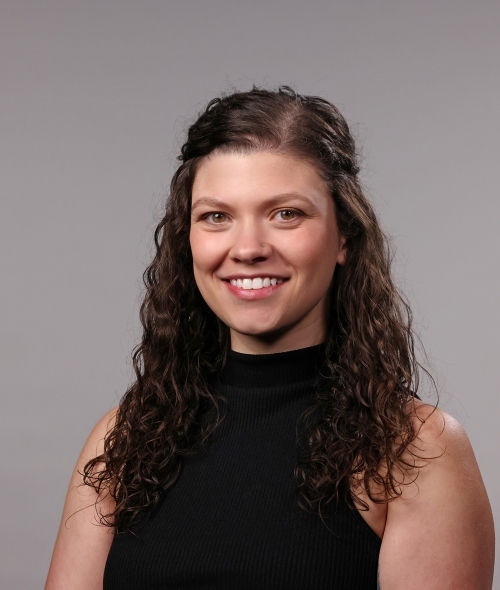 a woman with curly brown hair half-pinned back wearing a black turtleneck and smiling in front of a grey background