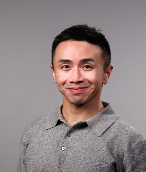a man with short dark hair wearing a grey collared shirt on a grey background