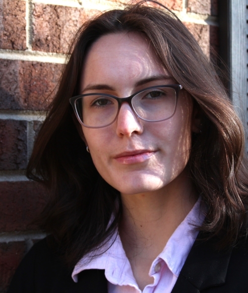 a young woman with dark brown hair wearing glasses and a collared shirt leaning against a brick wall