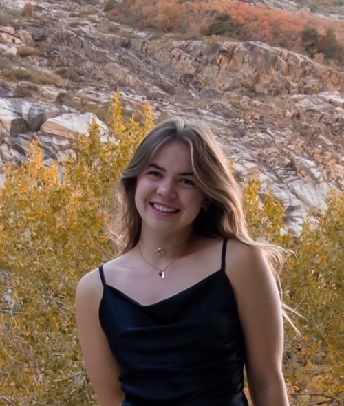 a young woman with long curly brown hair smiling wearing a black tank top in front of a rocky mountain